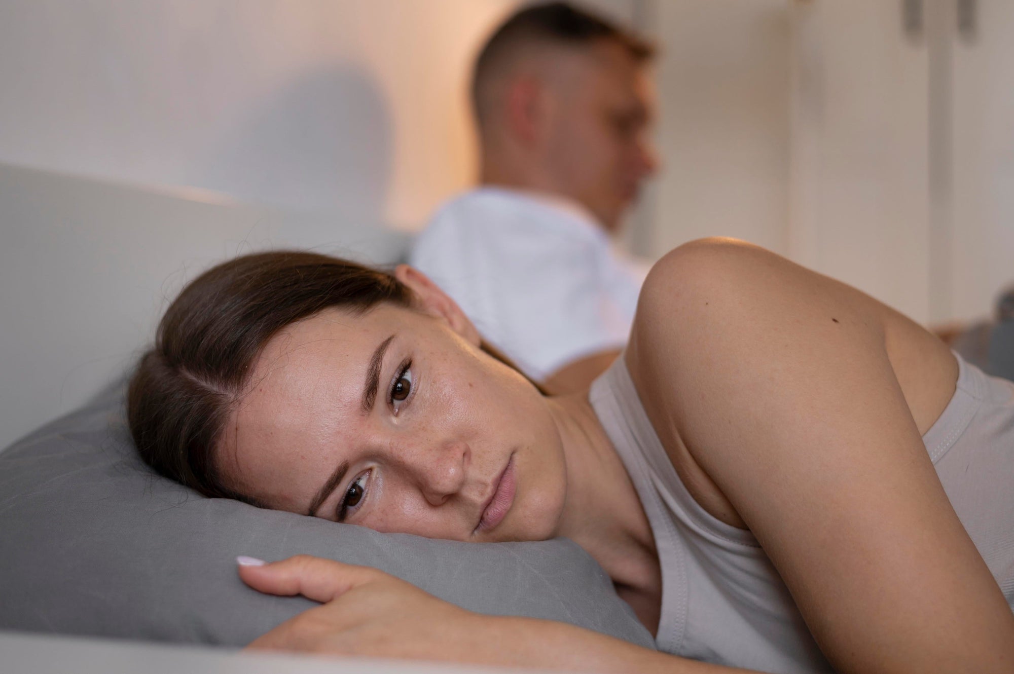 A woman lies awake in bed looking stressed while her partner sleeps soundly in the background.