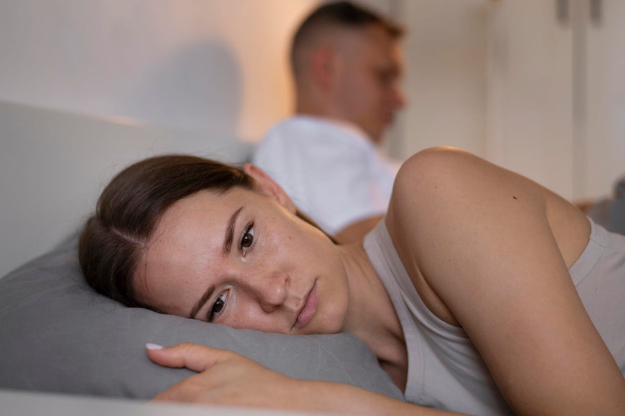A woman lies awake in bed looking stressed while her partner sleeps soundly in the background.