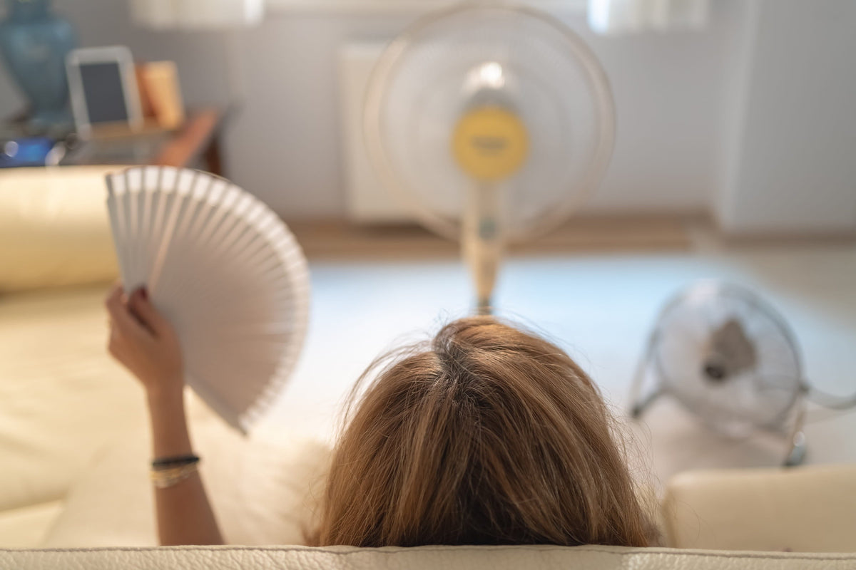 A woman with blonde hair holds a white hand fan while a standing electric fan blows air toward her.