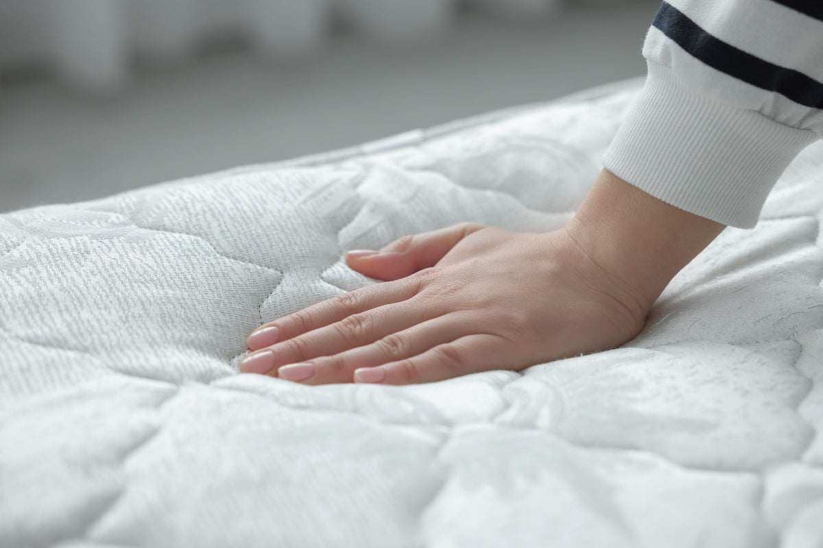 A person's hand presses firmly into a white, quilted mattress surface to test the softness.
