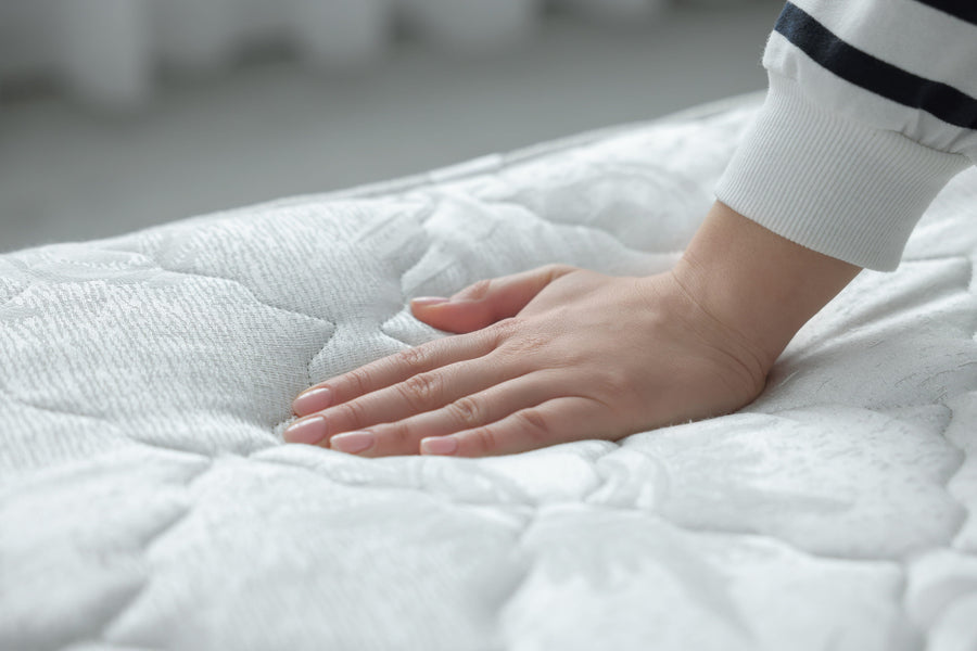 A person's hand presses firmly into a white, quilted mattress surface to test the softness.