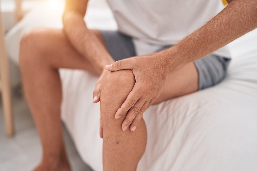 A person wearing a white shirt and grey shorts sits on the edge of a white bed, grasping their knee with both hands.