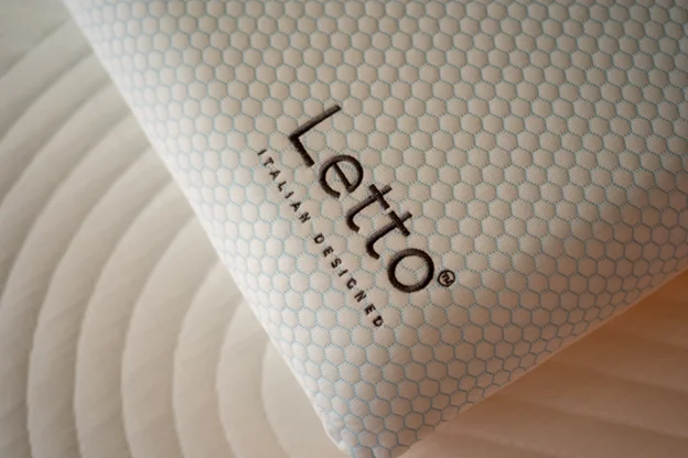 Close-up of a Letto pillow with a light blue hexagonal patterned cover, featuring the Letto logo and the words ‘Italian Designed,’ resting on a mattress with circular quilted stitching.