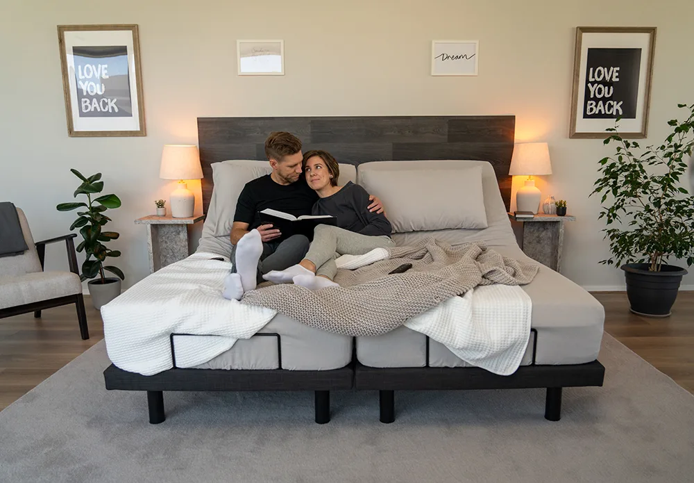A couple smiles while reading a book on a split adjustable bed, which can help find a comfortable position for how to sleep in the heat.