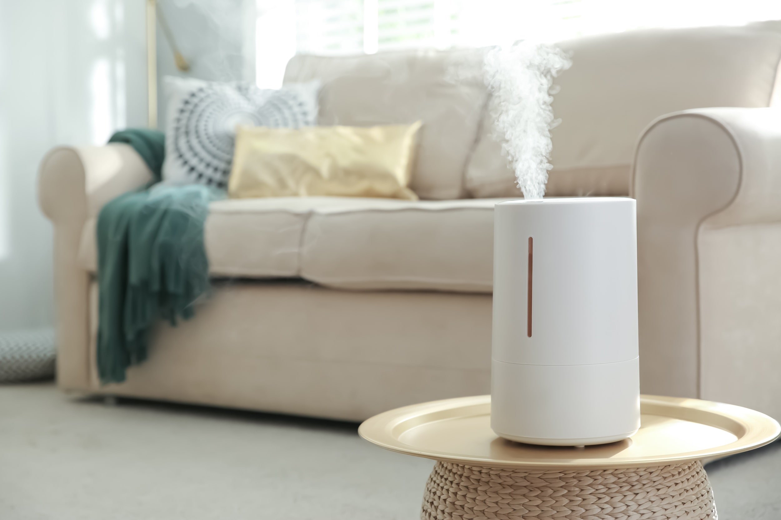 A white, cylindrical air purifier sits on a gold tray table in a living room, emitting a plume of white mist. In the blurred background is a beige sofa with decorative pillows and a throw blanket.