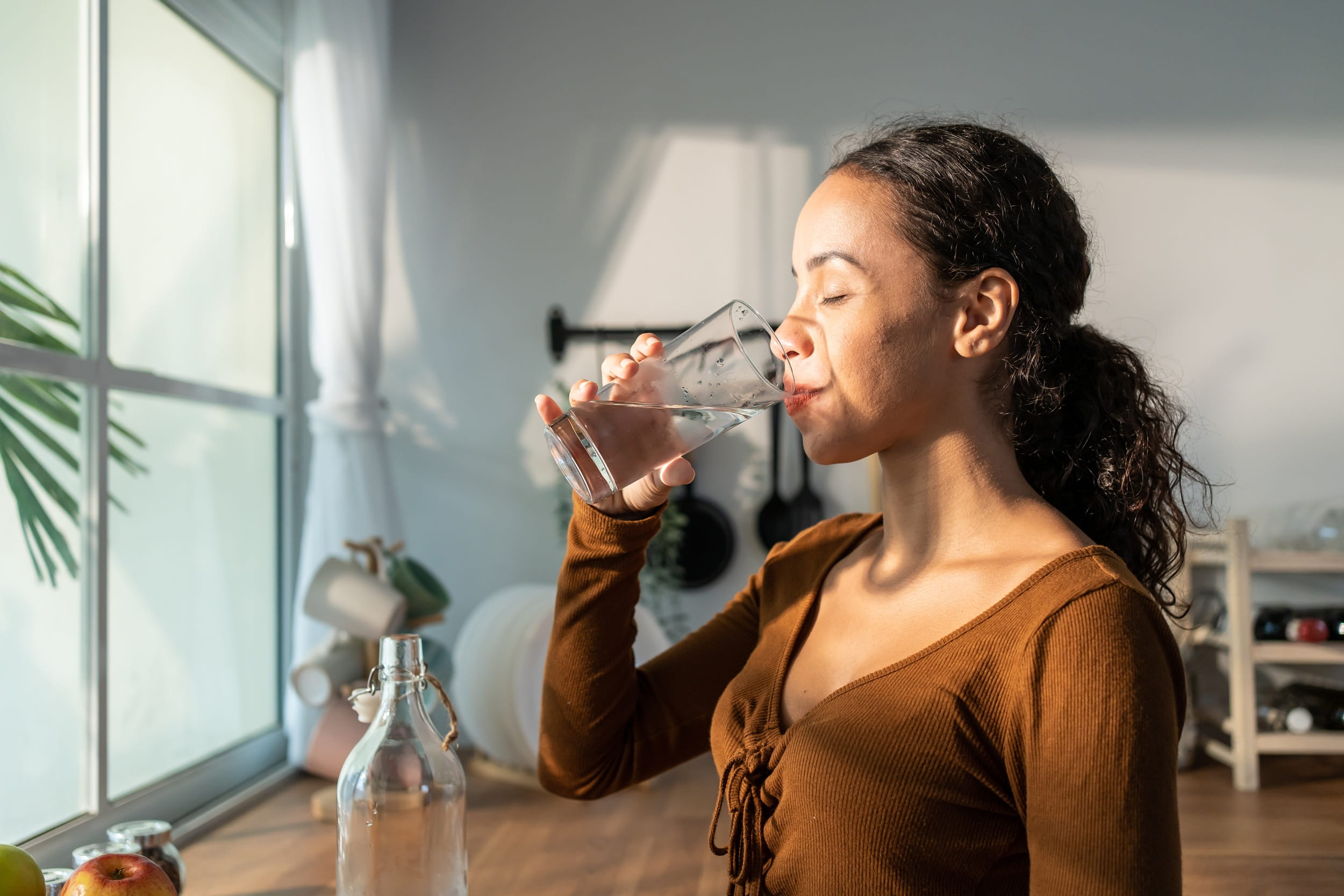 Woman drinking a glass of water in a sunlit kitchen, symbolizing hydration and wellness new year resolutions ideas.