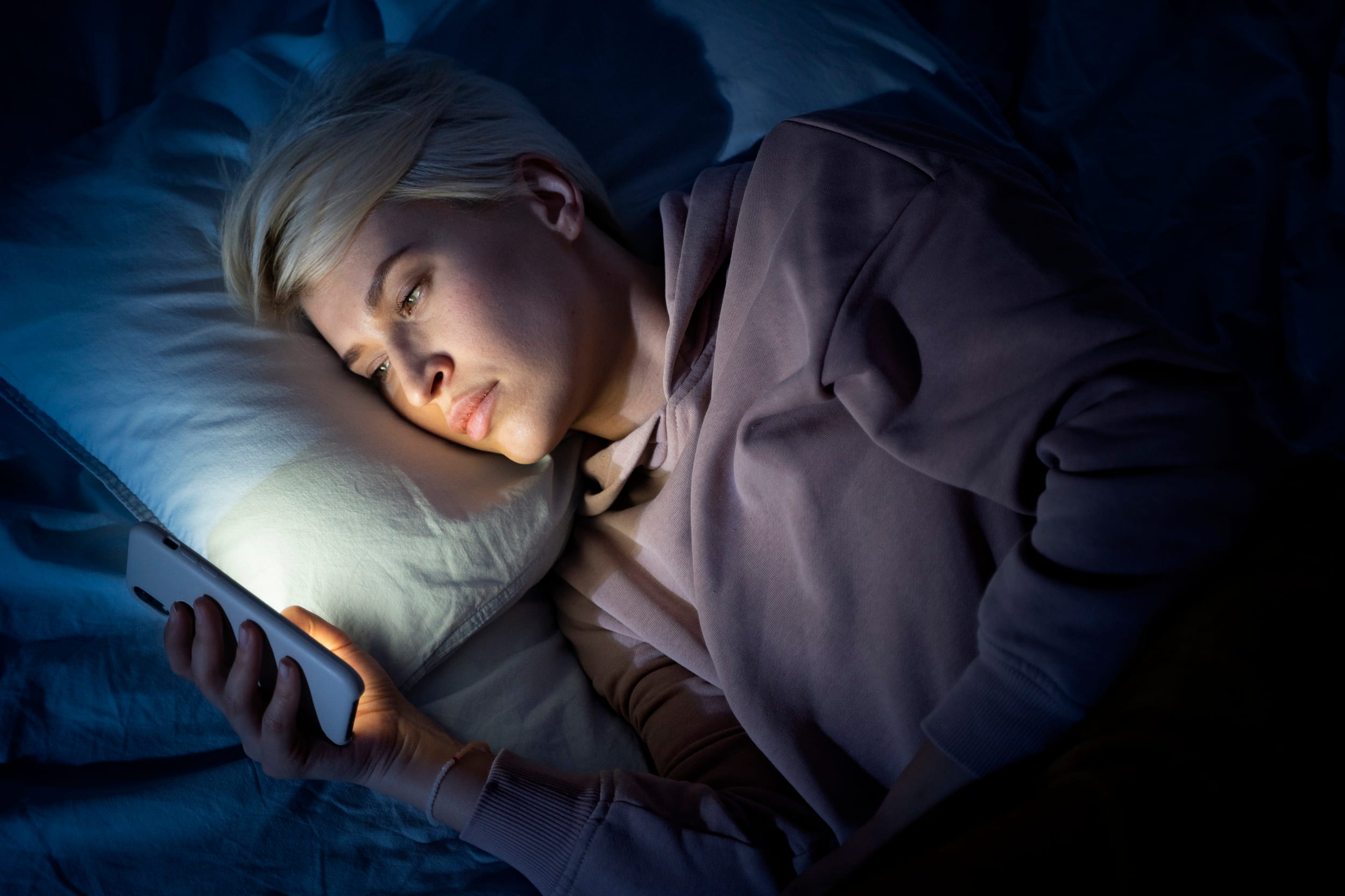 A woman lying in bed in the dark illuminated by her smartphone, highlighting habits that can disrupt restorative sleep