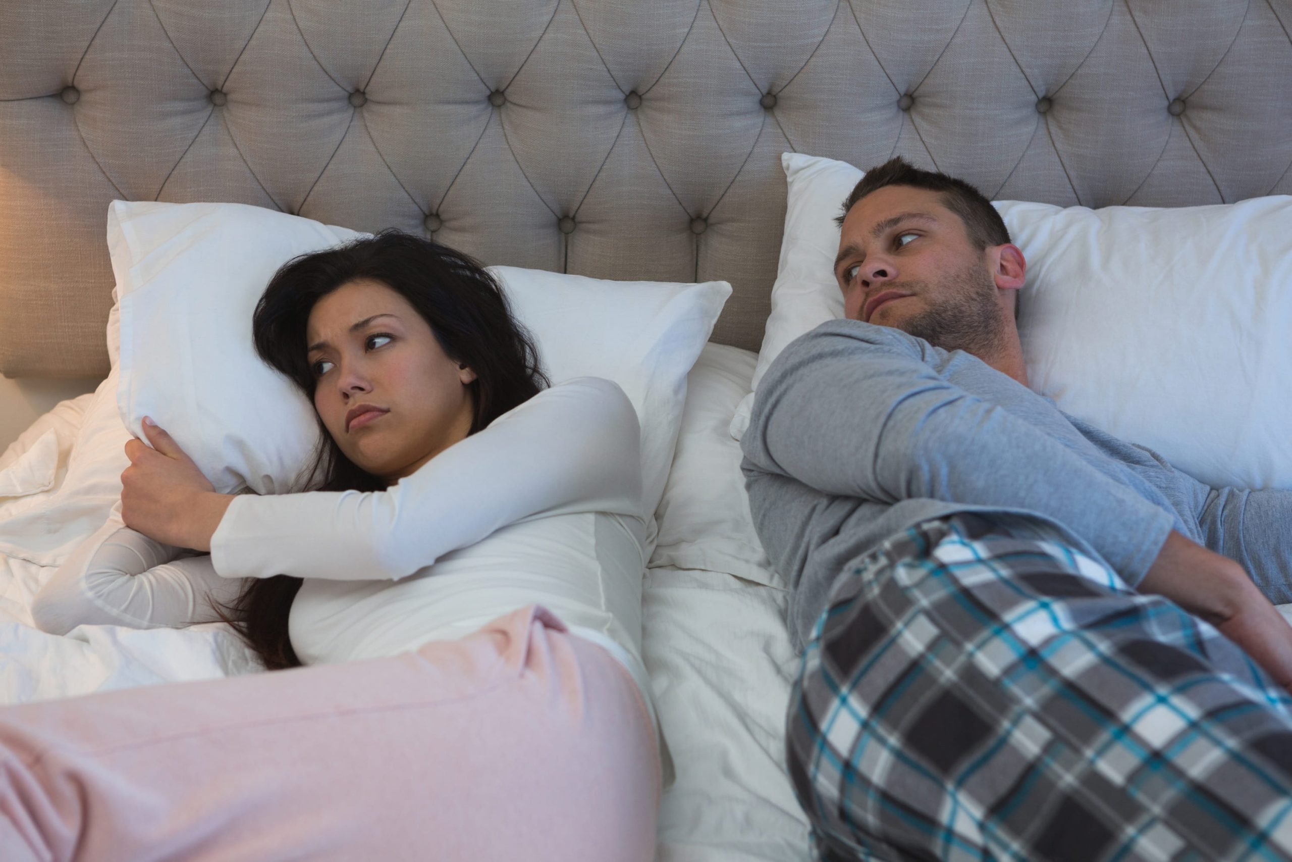 A frustrated woman lies awake clutching a pillow, facing away from her sleeping partner in a shared bed.
