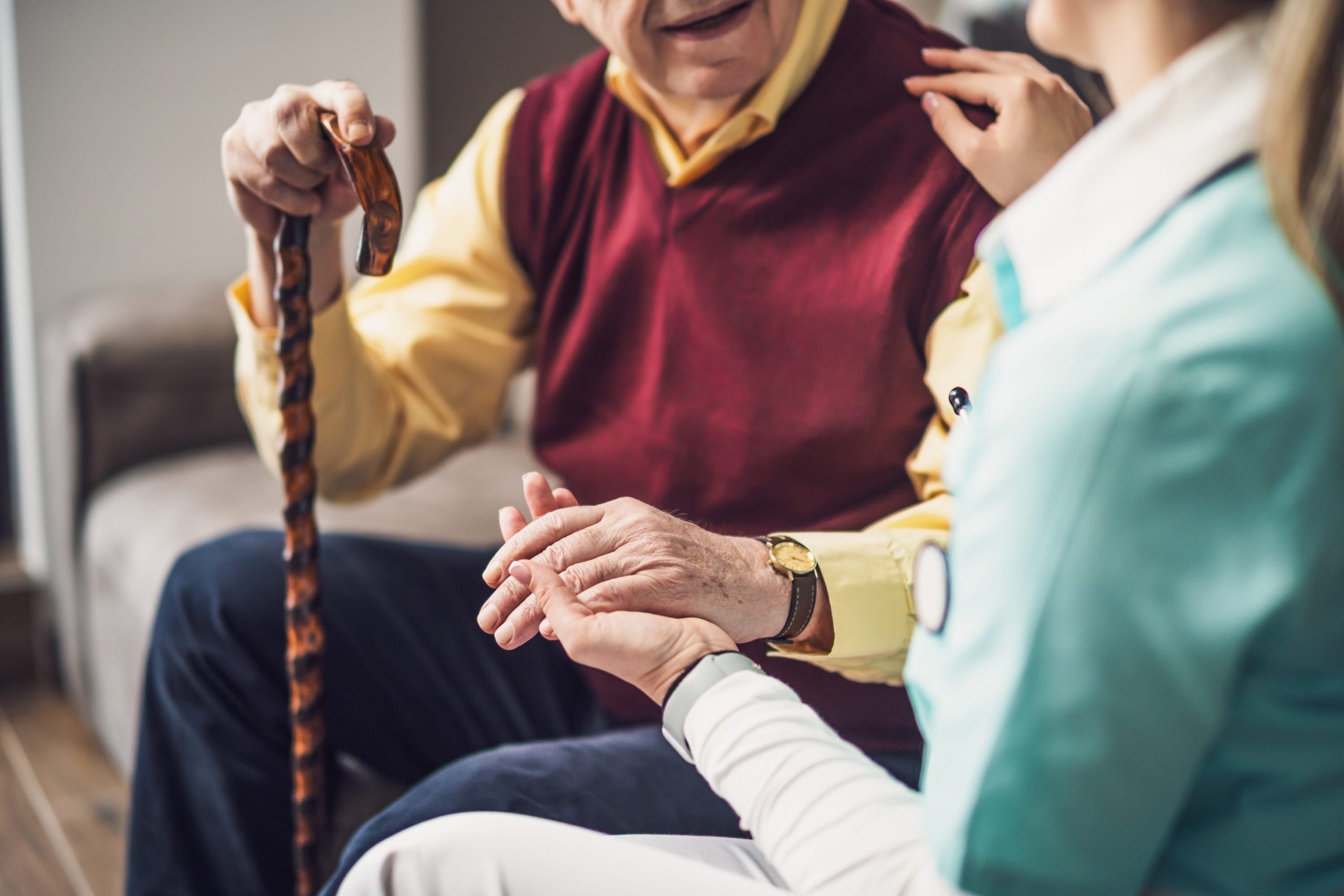 A close-up of a healthcare worker in a light blue uniform gently holding the hand of an elderly man who is holding a wooden walking cane.

