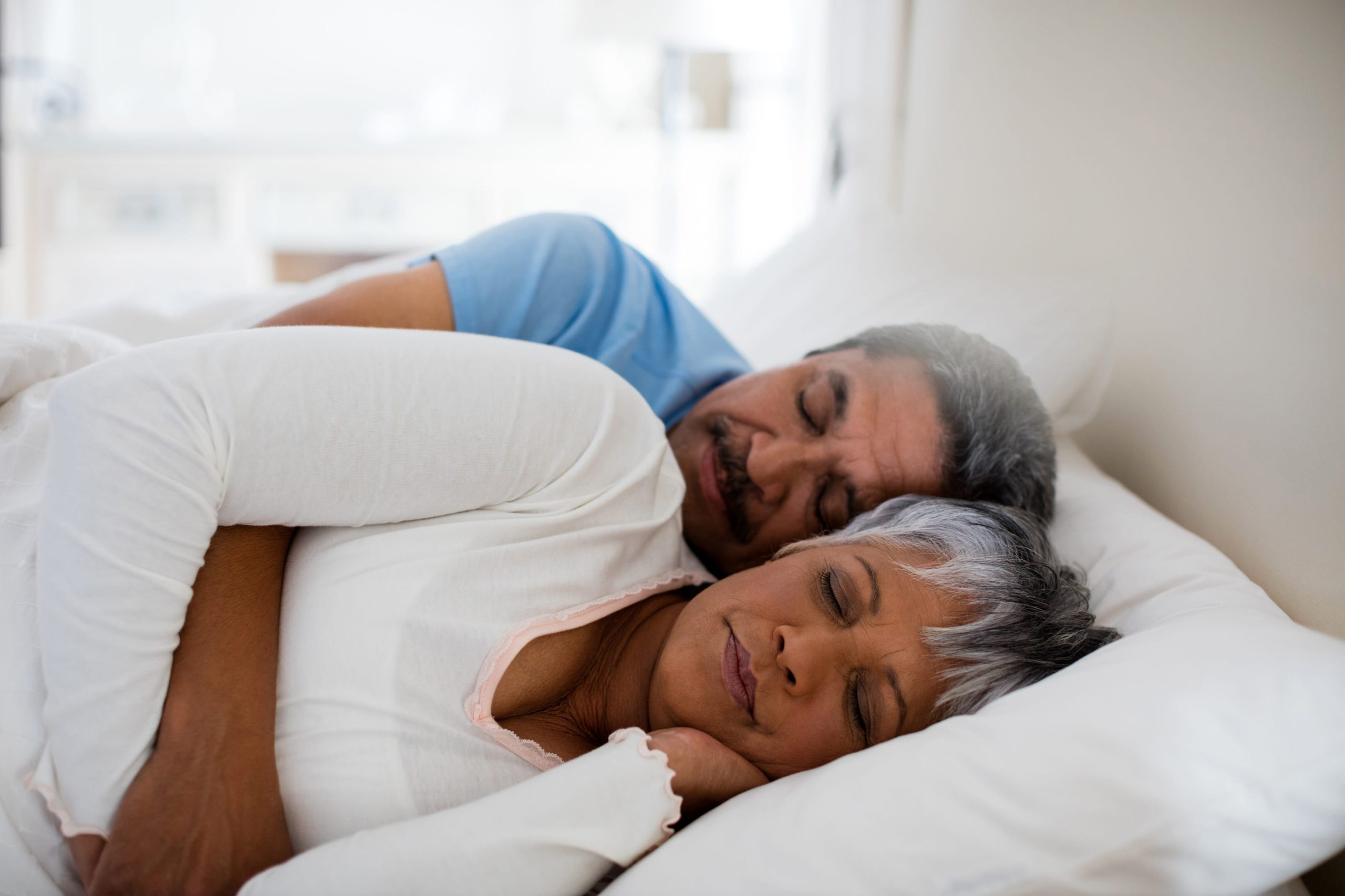 An older couple sleeping peacefully and deeply in a bed with white linens, highlighting the importance of restorative rest for brain health.
