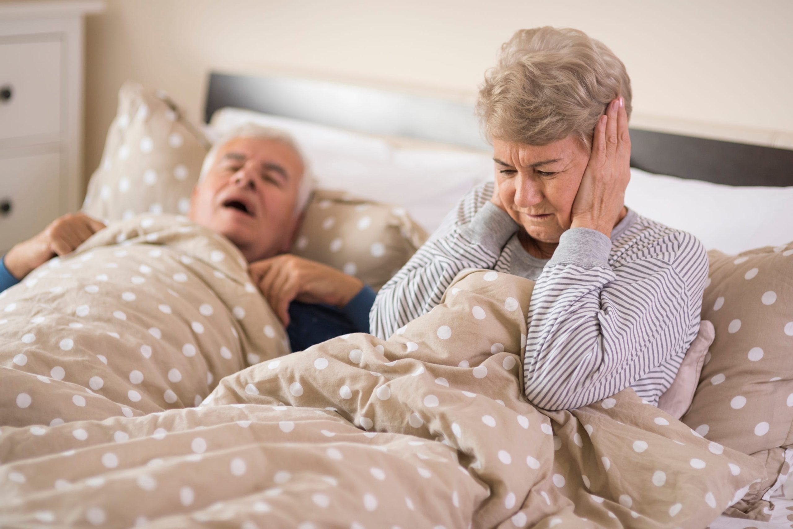 An older woman covers her ears in frustration to block out the noise of her partner snoring beside her.
