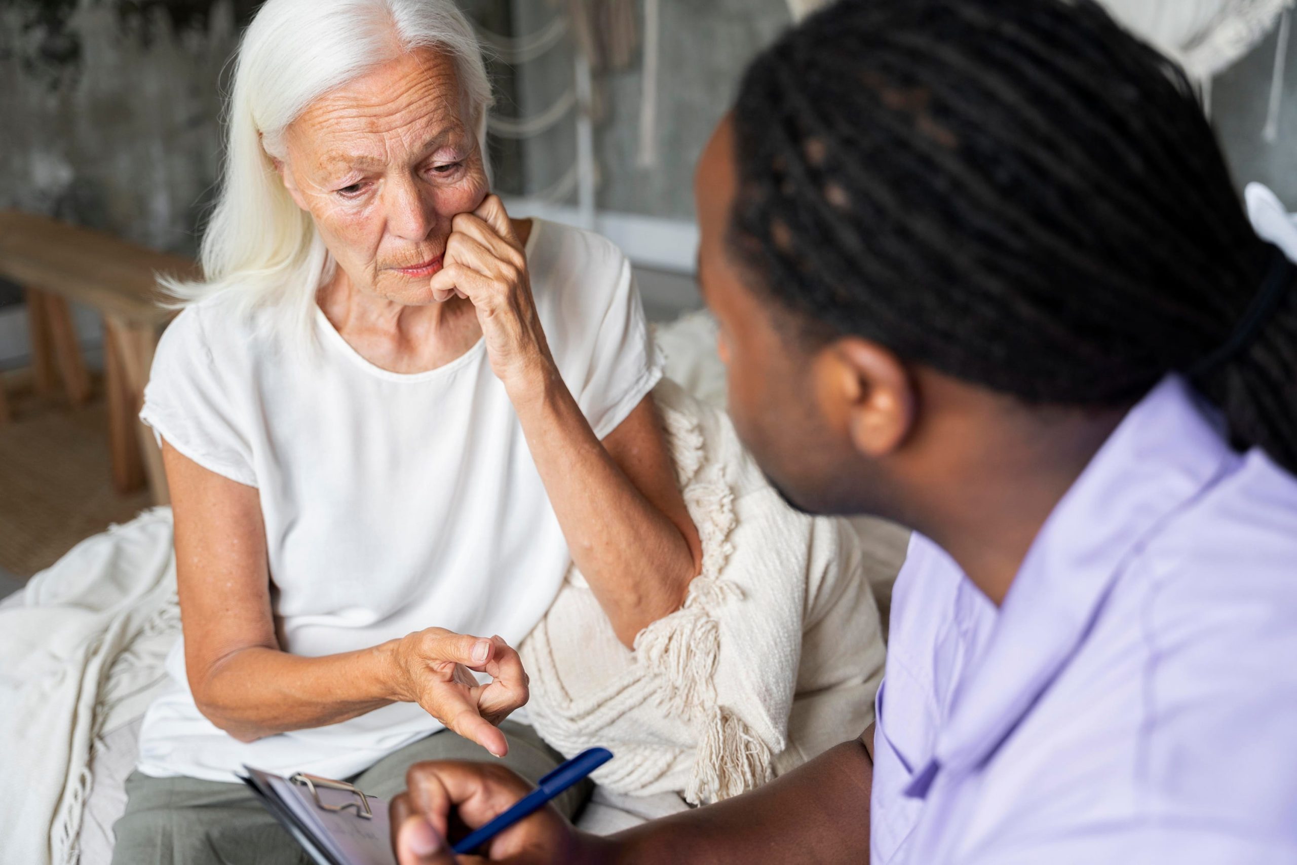 An older woman with white hair looking concerned while speaking with a healthcare professional who is taking notes on a clipboard in a home setting.