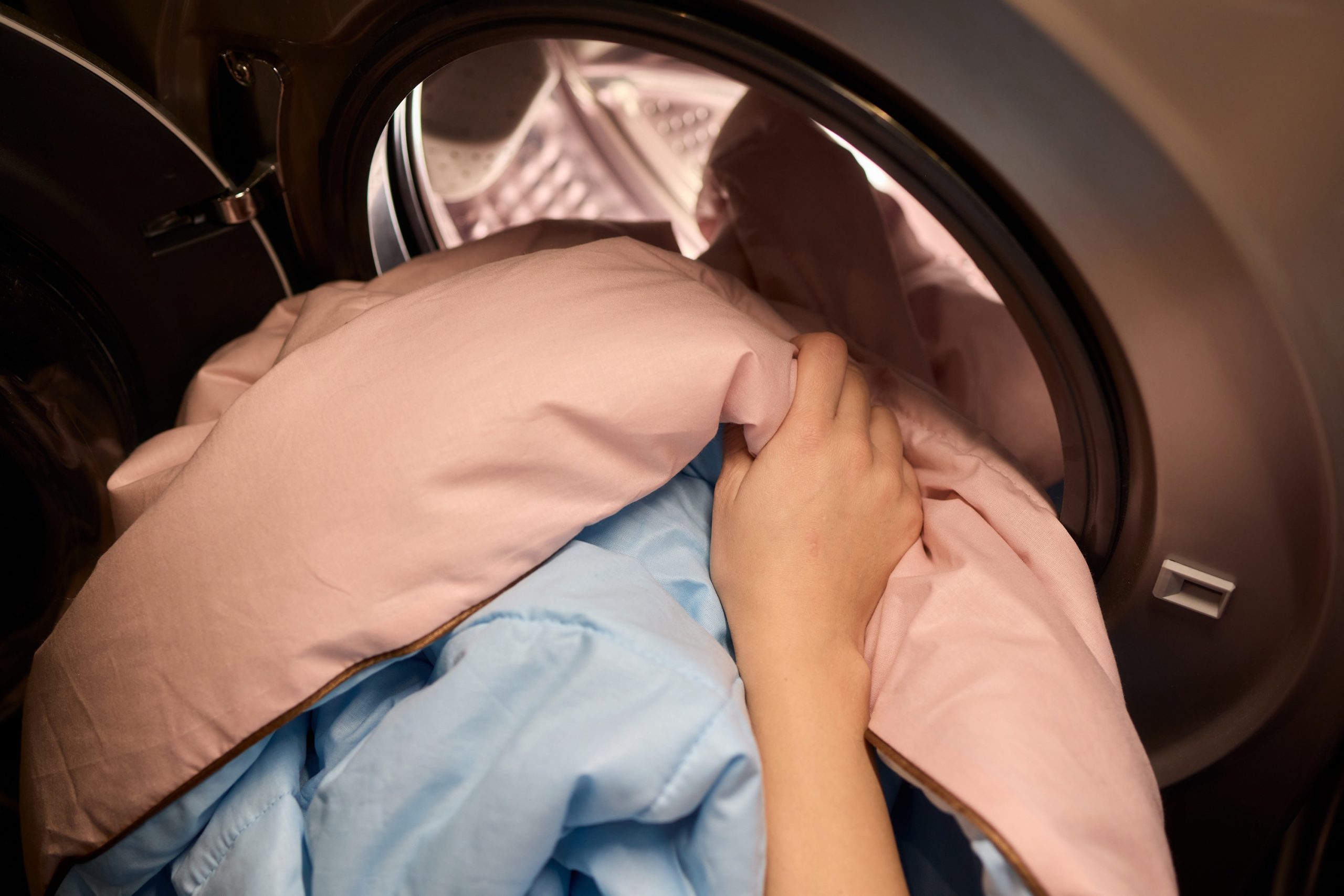Hands loading pink and blue sheets into a washing machine to remove bacteria and skin oils.