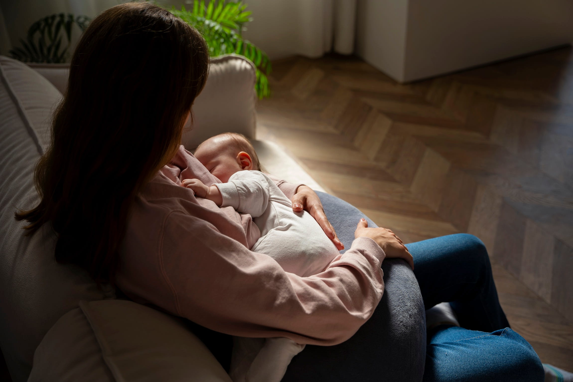 A mother sitting on a sofa in a dimly lit room, cradling her sleeping infant in a breastfeeding position, highlighting the physical demands and exhaustion of nighttime caregiving.
